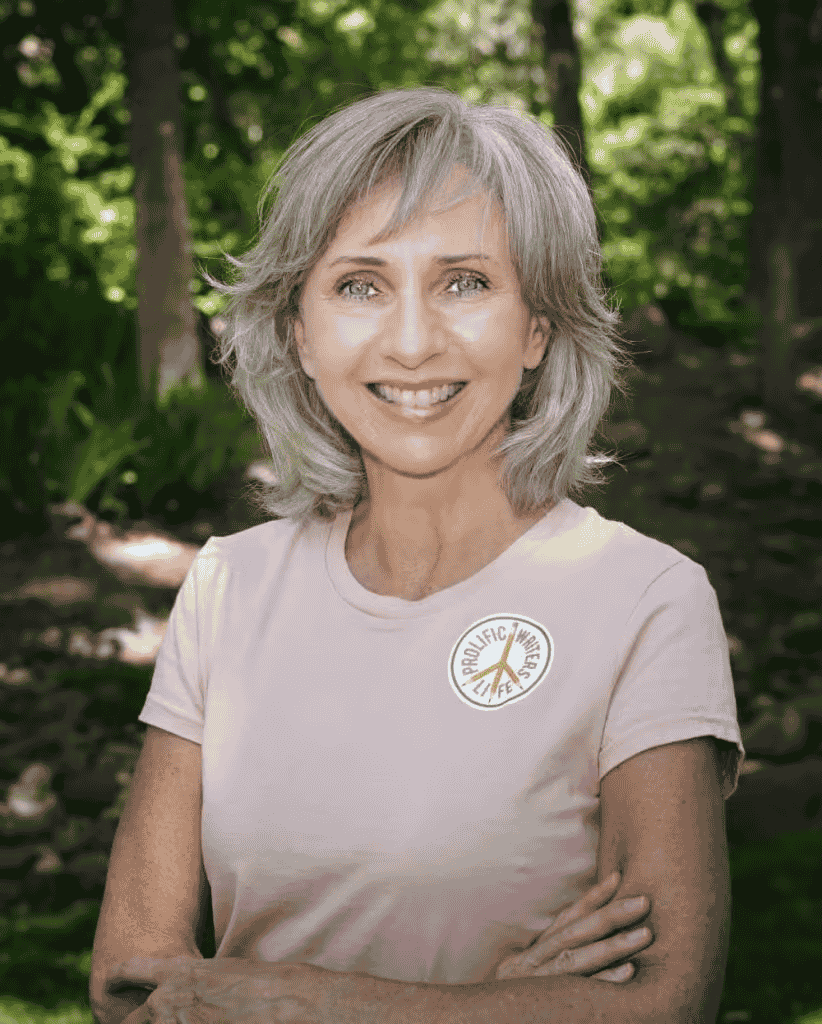 LorraineHaataiaPh_DProlificWritersLife - WC Vendors Lorraine Haataia, PhD wearing a pink shirt with the Prolific Writers logo with her arms crossed and standing in a forest